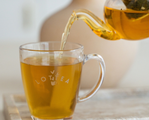 Een theeglas met daarop het Lottea logo. Het theeglas wordt ingeschonken met thee uit een theepot.