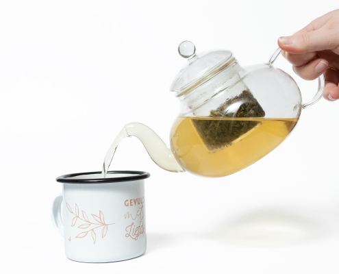 Een glazen theepot met daarin een filter waar losse thee in kan, er zit ook thee in en dit wordt ingeschonken in een theemok waar gevuld met liefde op staat.